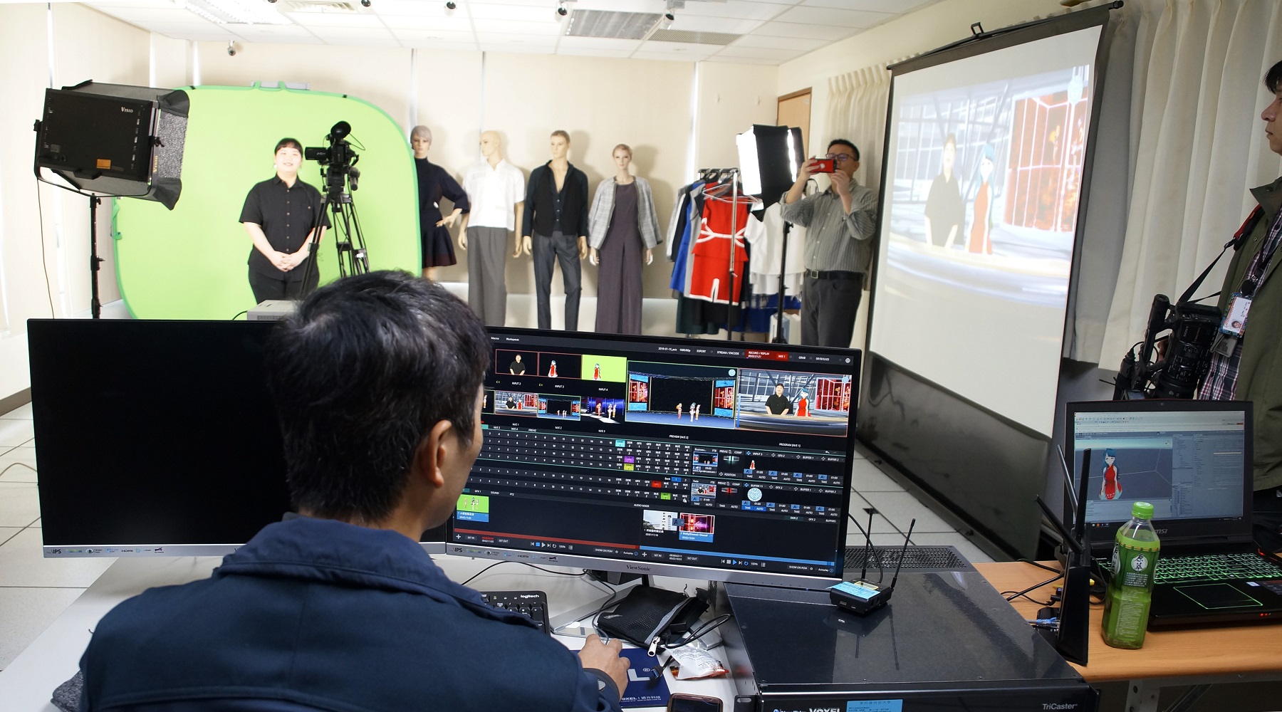 數媒實驗室能承接及輔導校內外數位教材開發與製播企劃。圖為實體主播（左，黑衣者）與虛擬主播（右邊投影螢幕紅衣者）合而為一，搭配虛擬背景，進行新聞播報！